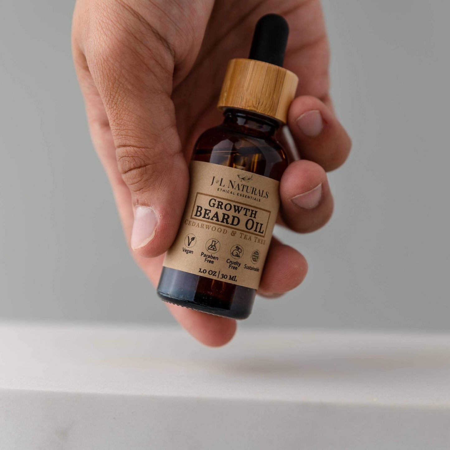 Hand holding J&L Naturals Growth Beard Oil with bamboo dropper top, shown against a minimalist grey background.