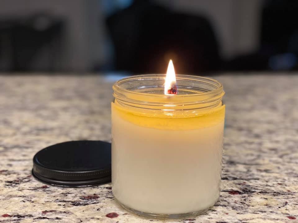 Lit soy candle in a clear glass jar with black lid resting nearby, placed on a speckled countertop with a softly blurred background.