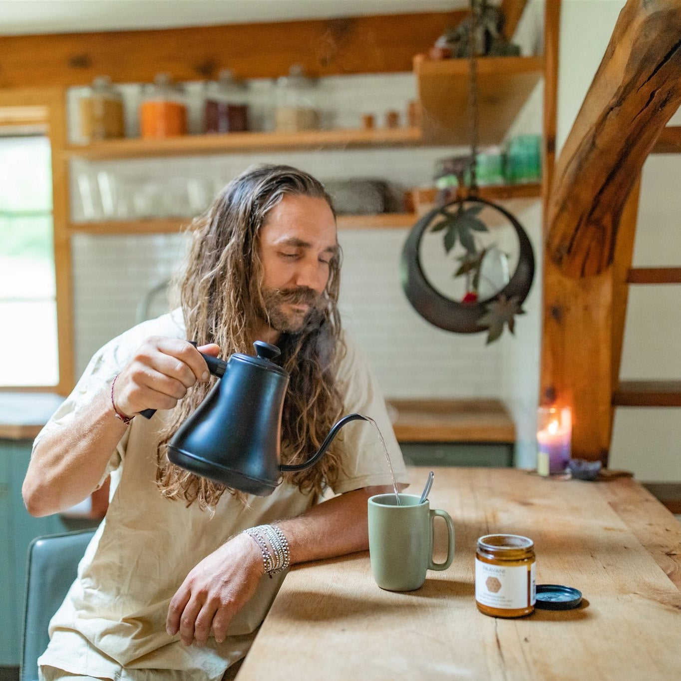 Man with long hair pouring hot water into a mug while preparing an Ashwagandha herbal tonic in a warm, rustic kitchen setting.