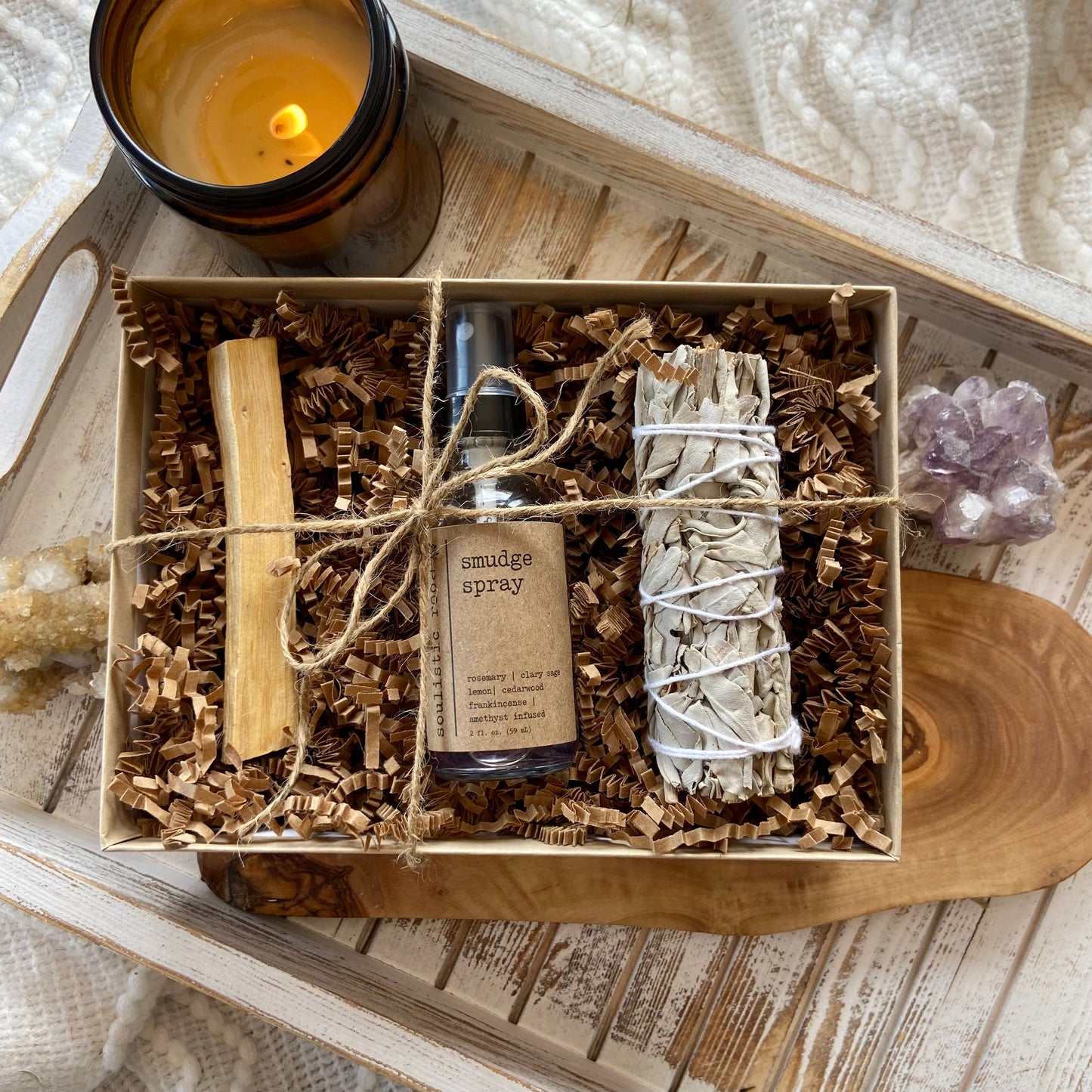 Top-down view of a Smudge & Cleanse Ritual Set including a white sage bundle, palo santo stick, and crystal-infused smudge spray in a gift box with shredded paper, displayed on a wooden tray with a lit candle and amethyst cluster.